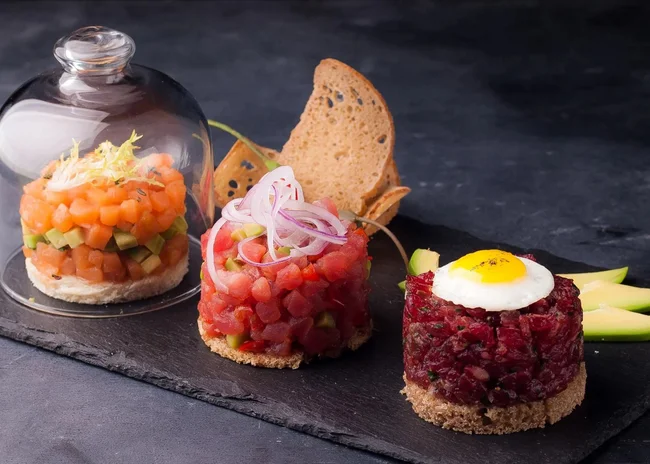 Der Salnerhof – das Hotel in Ischgl Drei kunstvoll angerichtete Tartare-Variationen mit Brot und Avocado auf schwarzem Stein