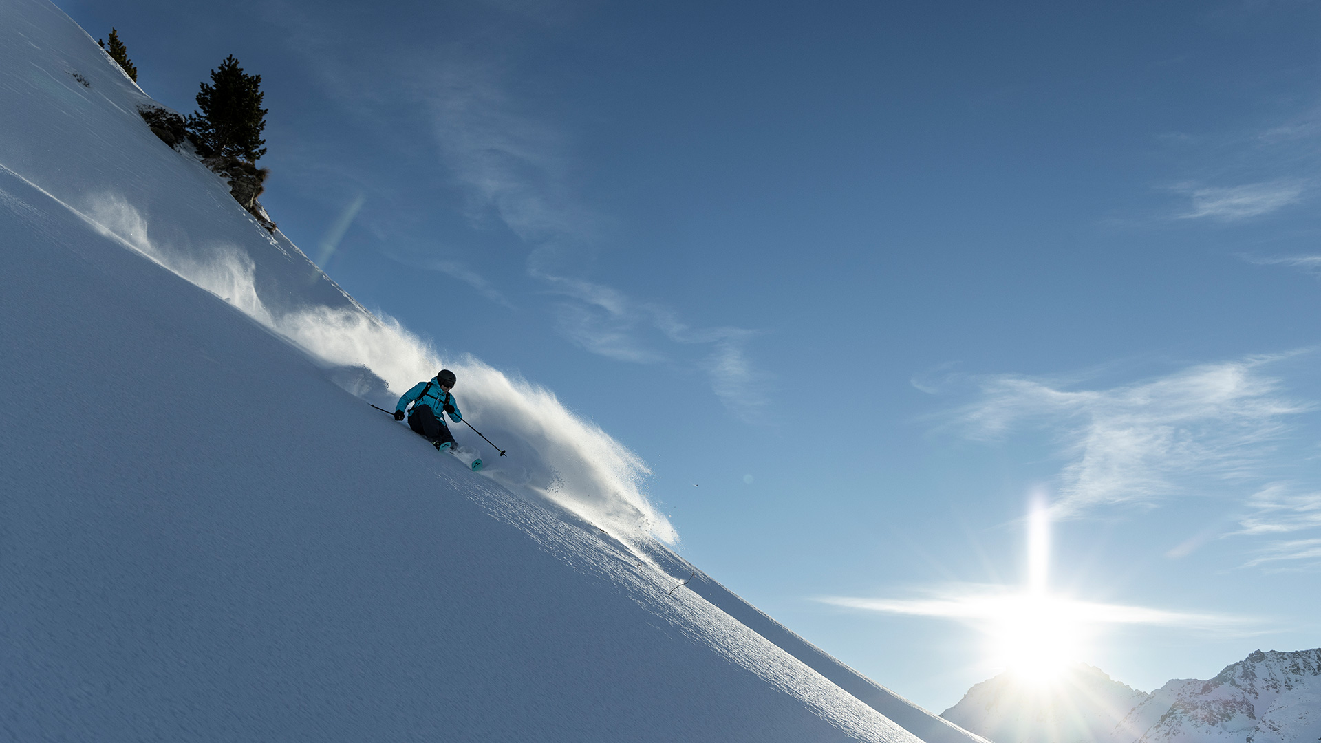 Skifahrer fährt bei Sonnenschein eine steile Tiefschneepiste hinab, aufgewirbelter Schnee und Bergpanorama im Hintergrund.