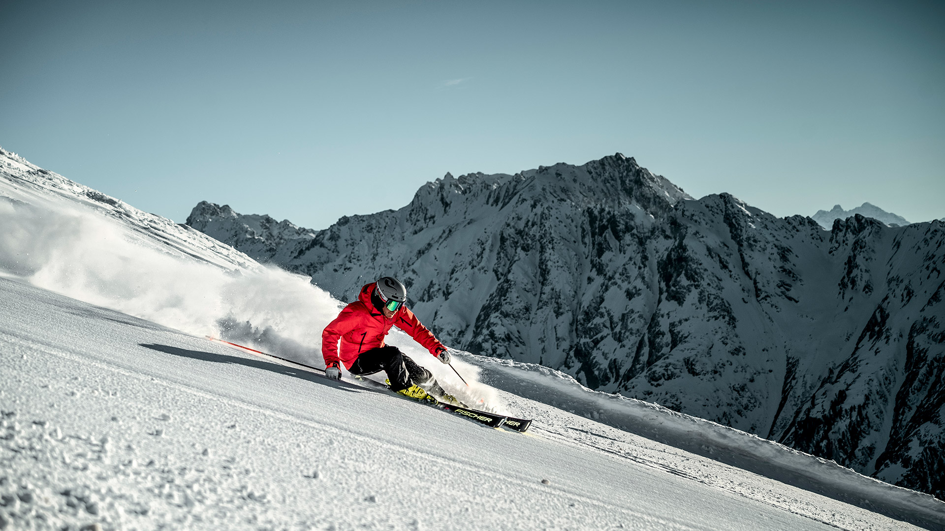 Skifahrer in roter Jacke carvt eine verschneite Piste hinab mit schneebedecktem Bergpanorama im Hintergrund.